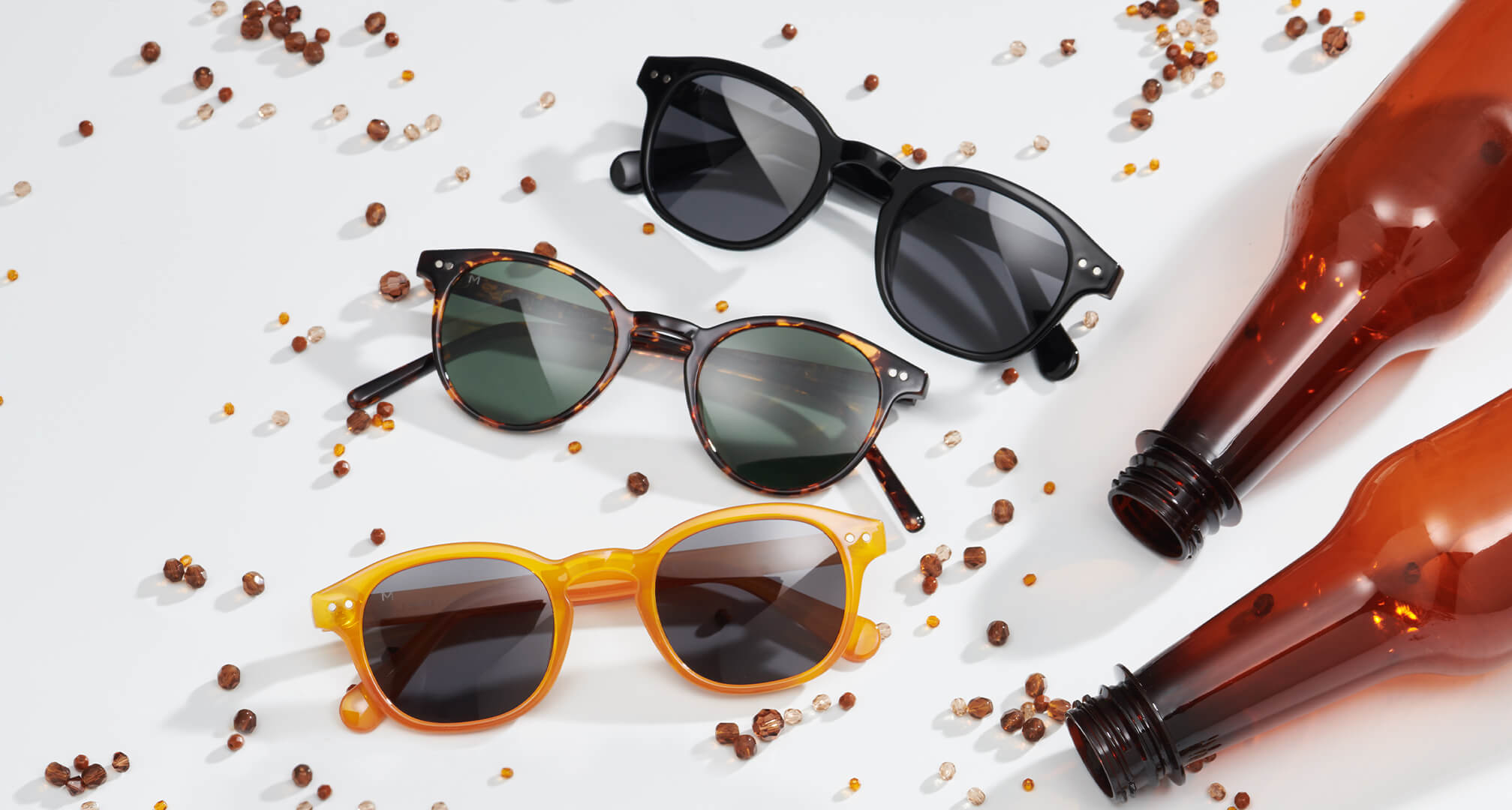 trije pari sončnih očal iz trajnostnih materialov na beli mizi, z okrasnimi kroglicami in plastičnimi steklenicami okoli njih